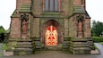 A historic church entrance with a red and gold stained glass window, flanked by statues and intricate stone carvings.