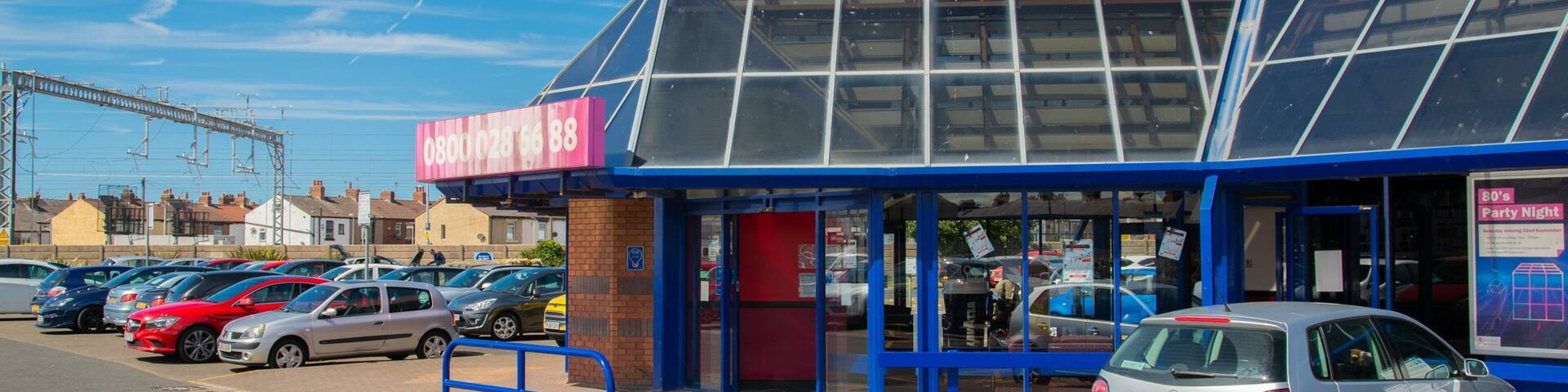 A parking lot with several cars, a building with a blue roof and a large glass window, and a pink sign with a phone number.