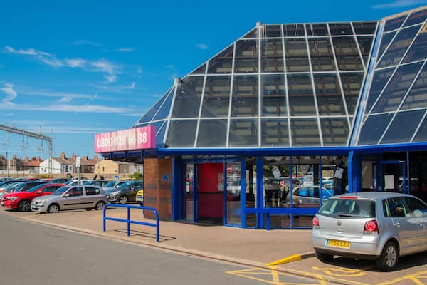Un parking avec plusieurs voitures, un bâtiment doté d’un toit bleu et d’une grande fenêtre en verre, et une enseigne rose avec un numéro de téléphone.