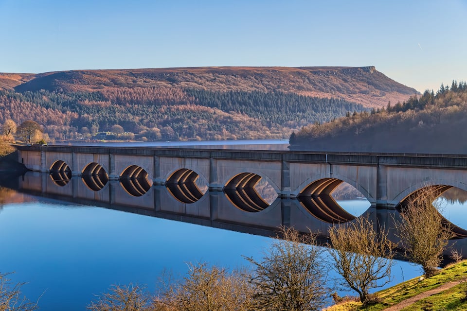 A bridge with unique arch designs over a calm lake, surrounded by forested hills.