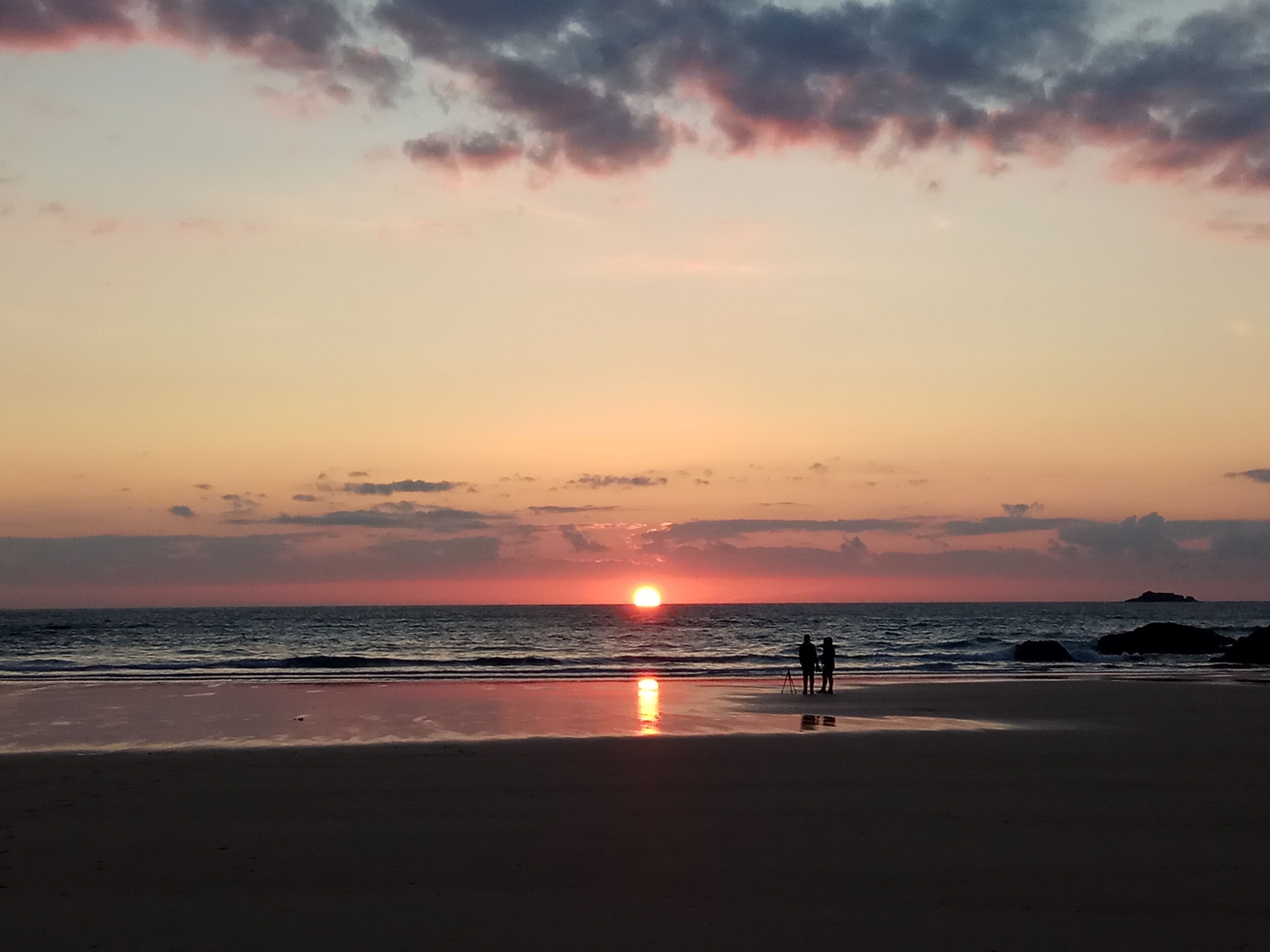 A sunset over a beach with two people silhouetted against the sky.