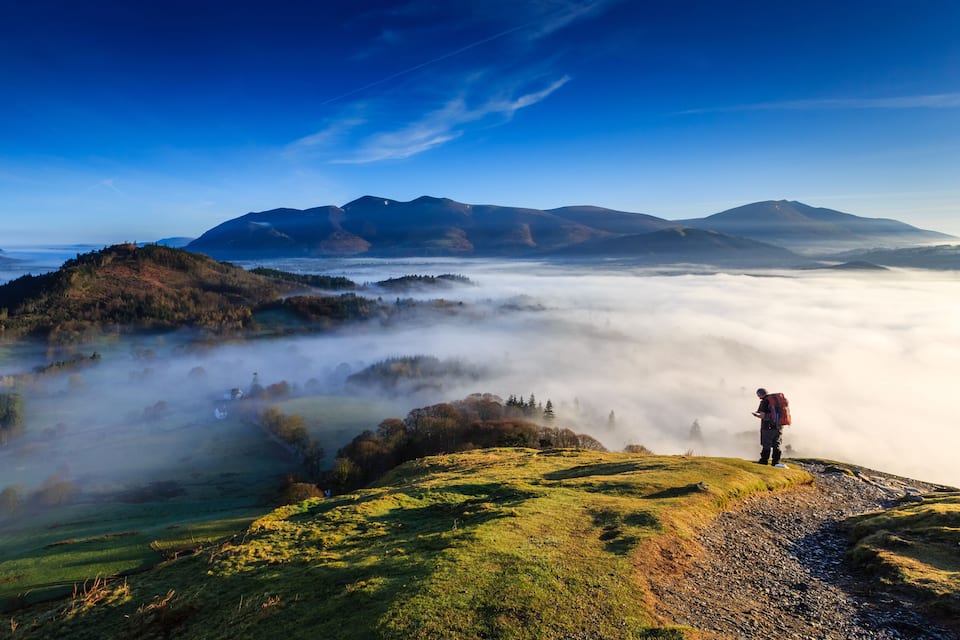 A hiker on a grassy hill overlooking a valley shrouded in mist, with mountains in the background.