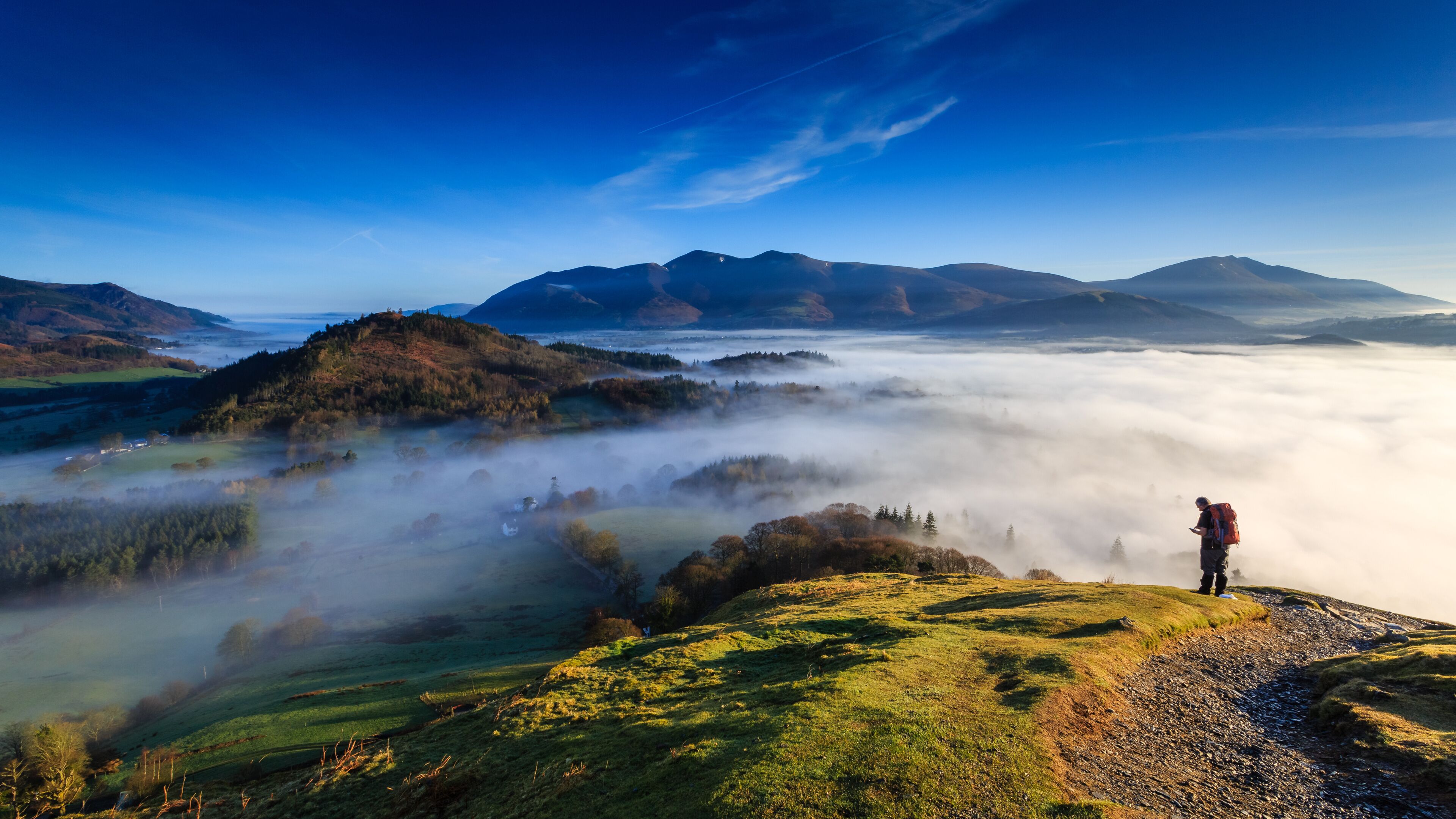 A hiker on a grassy hill overlooking a valley shrouded in mist, with mountains in the background.