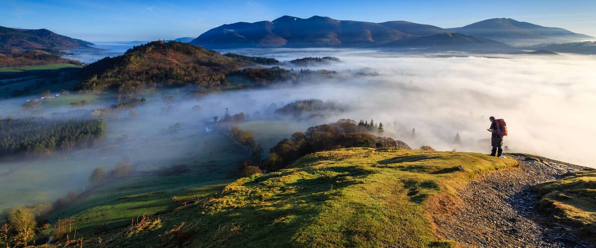 A hiker on a grassy hill overlooking a valley shrouded in mist, with mountains in the background.