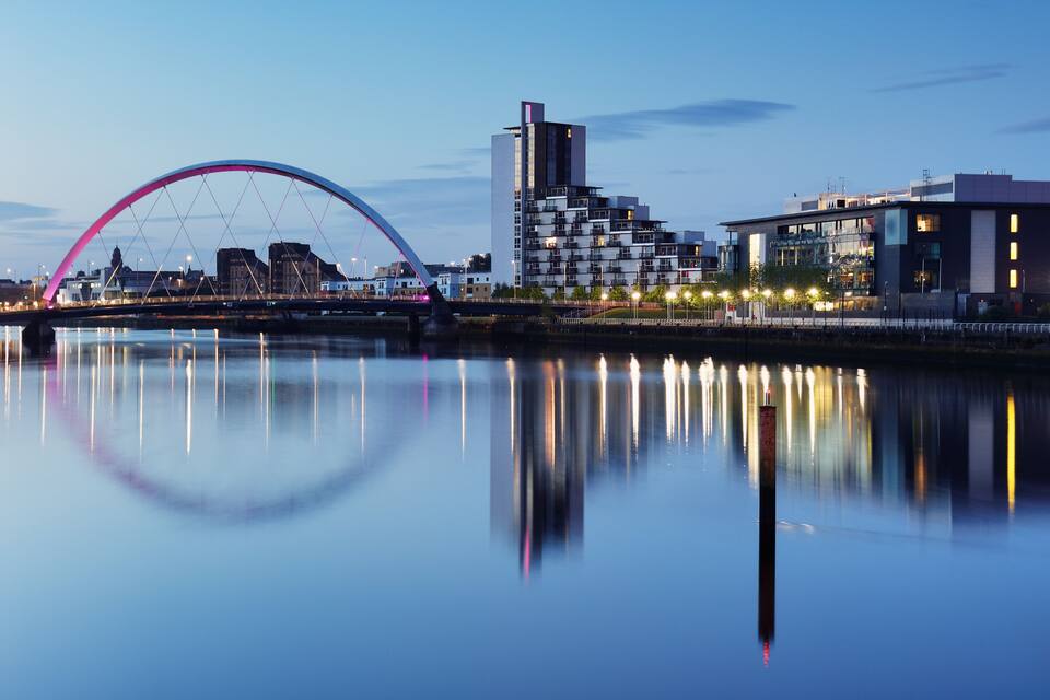 A cityscape with a modern bridge, a tall building, and a calm river reflecting the structures.