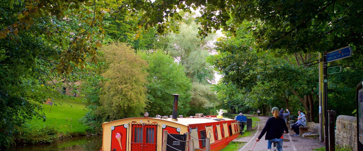 A narrow canal with a red and yellow boat docked on the left, a paved path with people walking and a dog, and lush green trees lining the path.