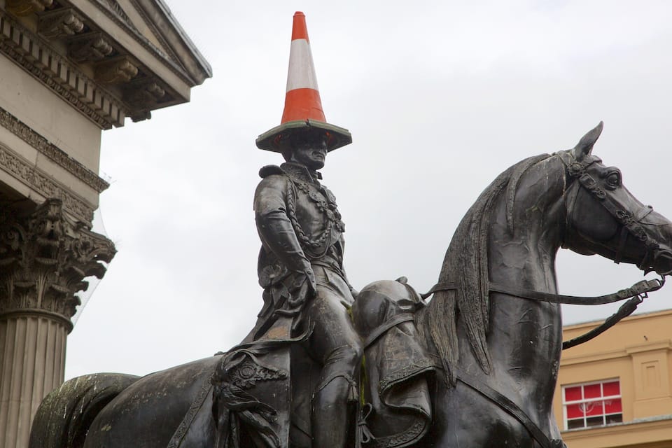 A statue of a mounted figure with a traffic cone on their head, set against a building with classical architecture.