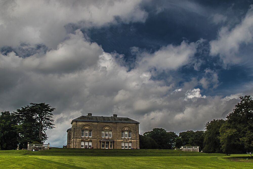 A large, historic building with a symmetrical facade and arched windows, surrounded by a well-maintained lawn and trees, under a cloudy sky.