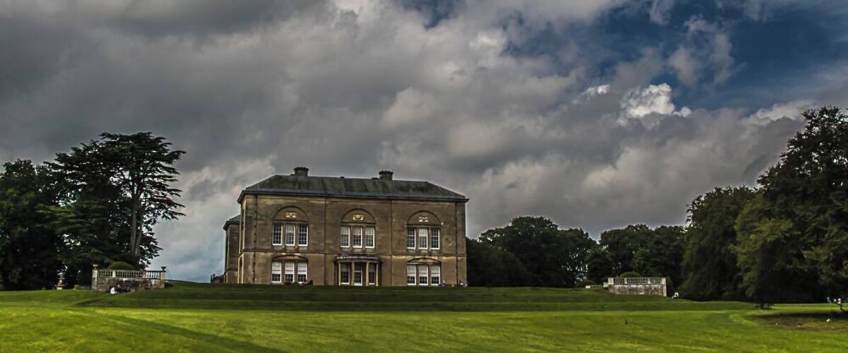 A large, historic building with a symmetrical facade and arched windows, surrounded by a well-maintained lawn and trees, under a cloudy sky.