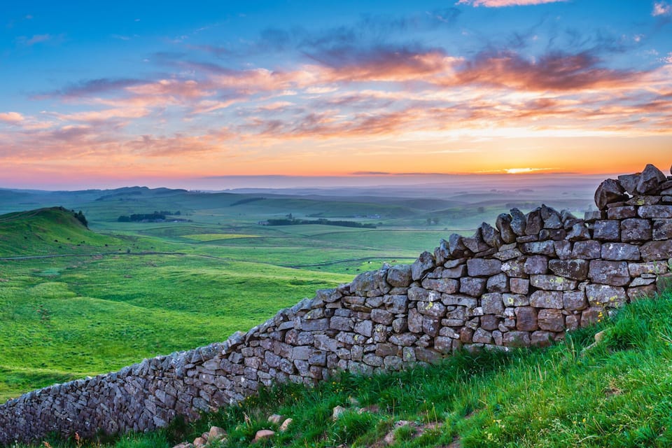 Een stenen muur, glooiende heuvels en een zonsondergang boven een groen landschap.