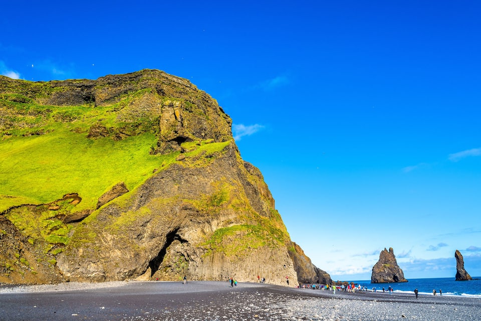 A coastal landscape with a prominent green hill, a rocky cliff, and distinctive rock formations in the sea.