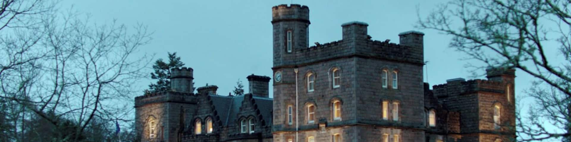 A large, historic stone building with multiple towers and arched windows, surrounded by trees and a well-maintained lawn.
