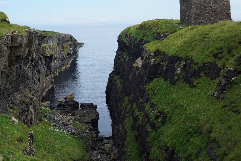 A coastal landscape with a narrow rocky passage, grassy cliffs, and a stone ruin.