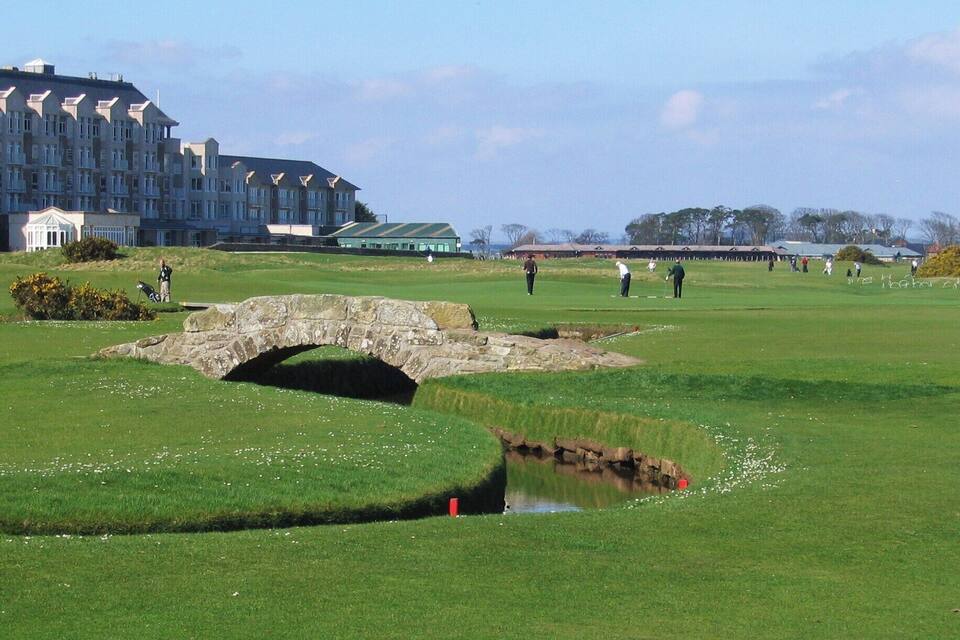 A golf course with a stone bridge over a small stream, surrounded by lush green grass and a large building in the background.