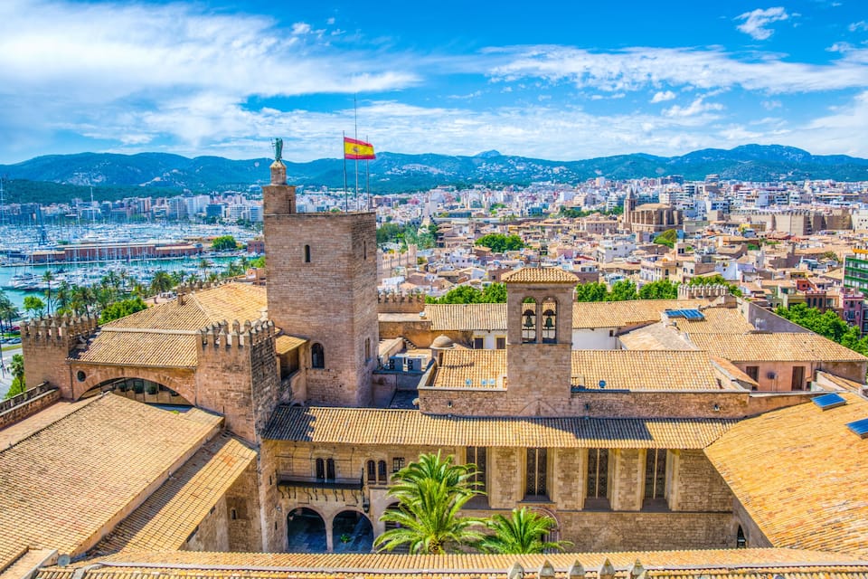 A historic cityscape with a prominent tower and a flag, surrounded by a harbor and mountains in the background.