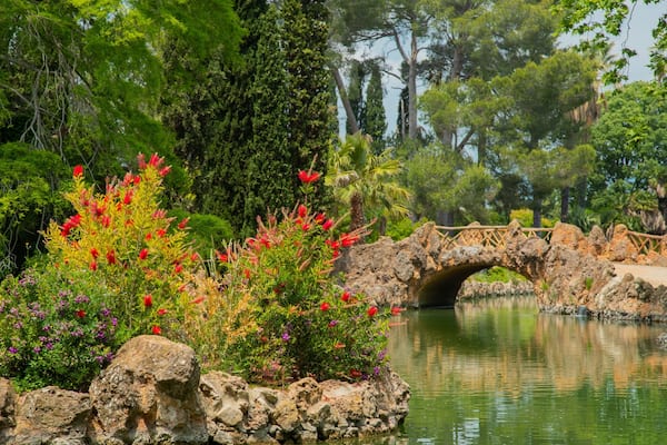 Un pont de pierre enjambant un étang paisible, entouré d’une végétation luxuriante et de fleurs aux couleurs vives.