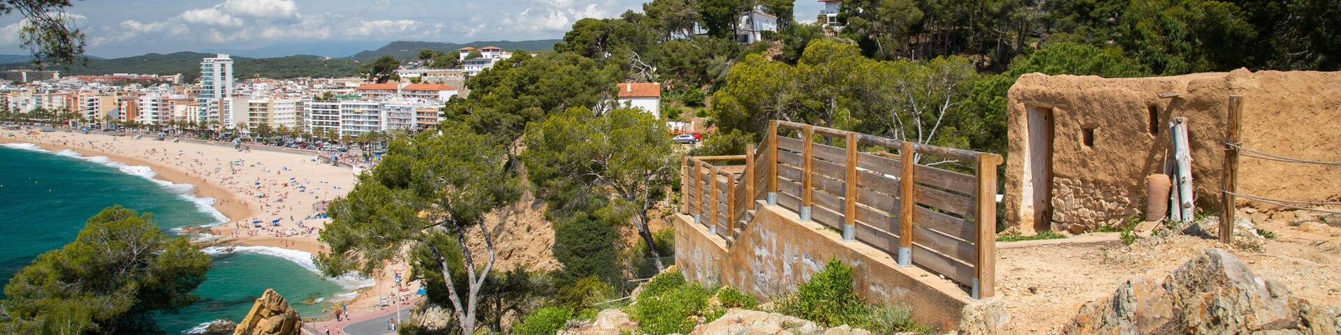 Paisaje costero con un acantilado rocoso, una escalera de madera, una playa y una ciudad al fondo.