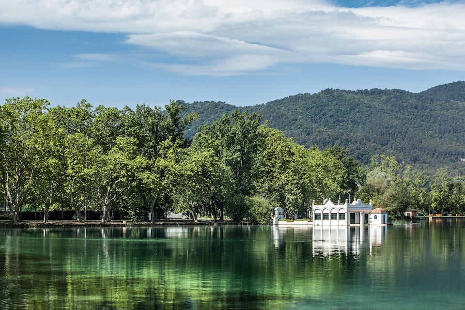 A serene lake with a small house on stilts, surrounded by lush green trees and hills in the background.