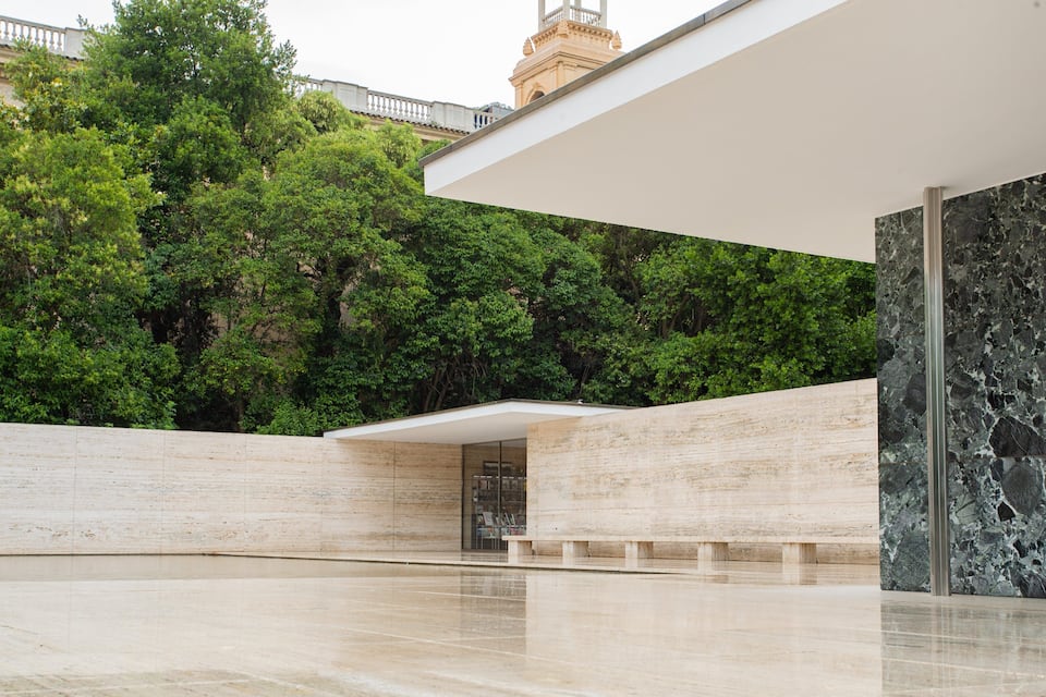 Um edifício moderno com entrada de vidro, rodeado por árvores verdes exuberantes e uma parede de mármore.