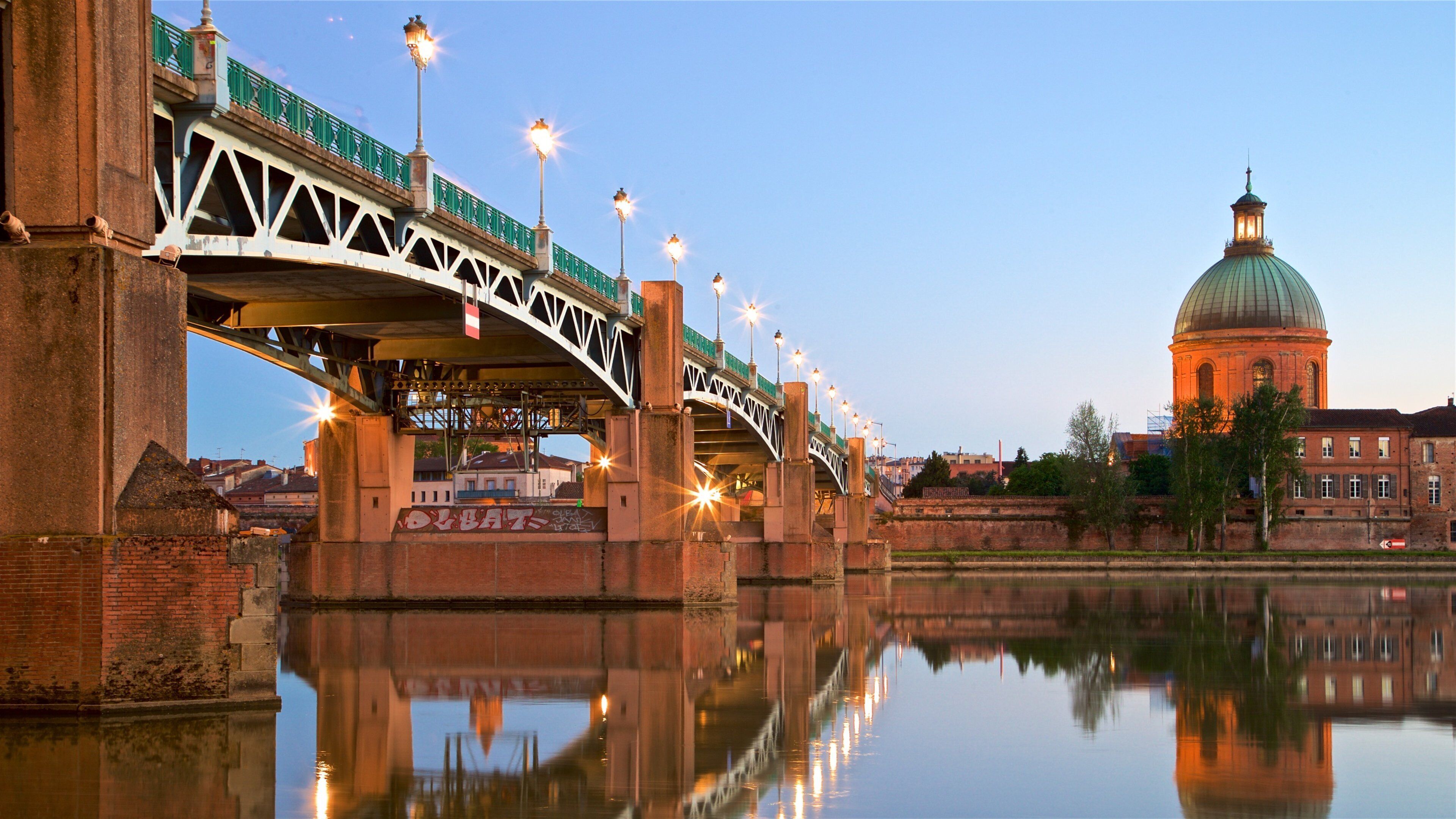 A bridge with street lamps over a calm river, reflecting the structure and a domed building in the background.
