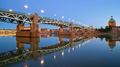 Un puente con luces que se reflejan en el agua, un edificio de forma abovedada y un río tranquilo.