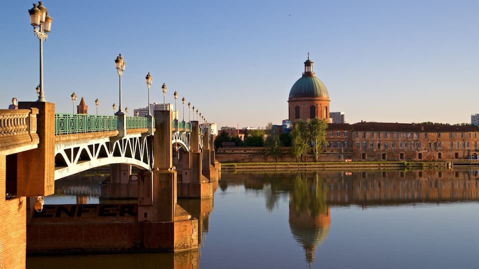A bridge with ornate street lamps, a calm river reflecting the buildings, and a domed building in the background.