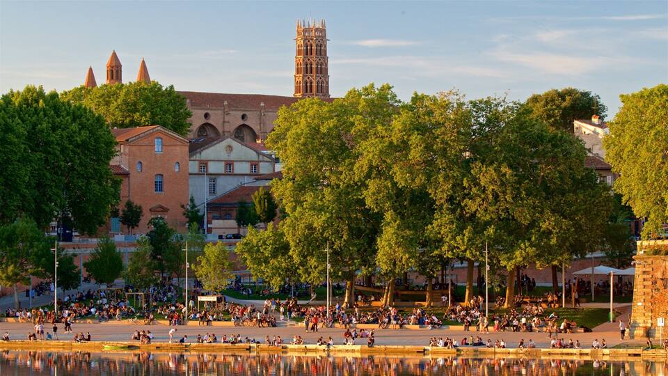 A riverside park with people relaxing, a historic church, and a tall tower in the background.