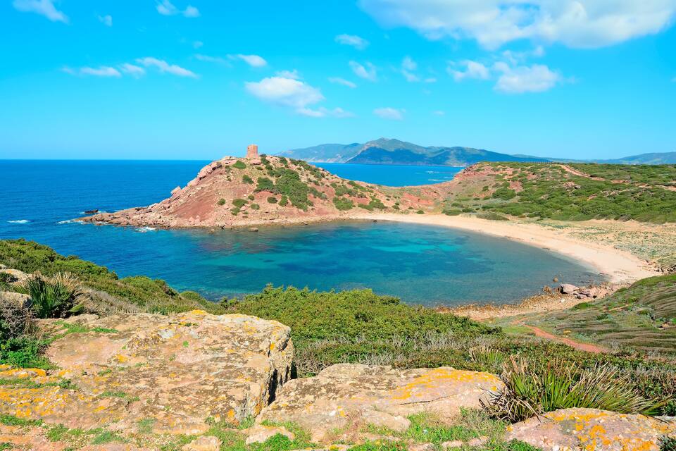 A coastal landscape with a clear blue sea, rocky terrain, and a small hill with a tower.