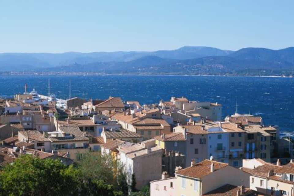 Uma cidade costeira com uma torre imponente, edifícios em terraços e vista clara para o mar e montanhas ao longe.