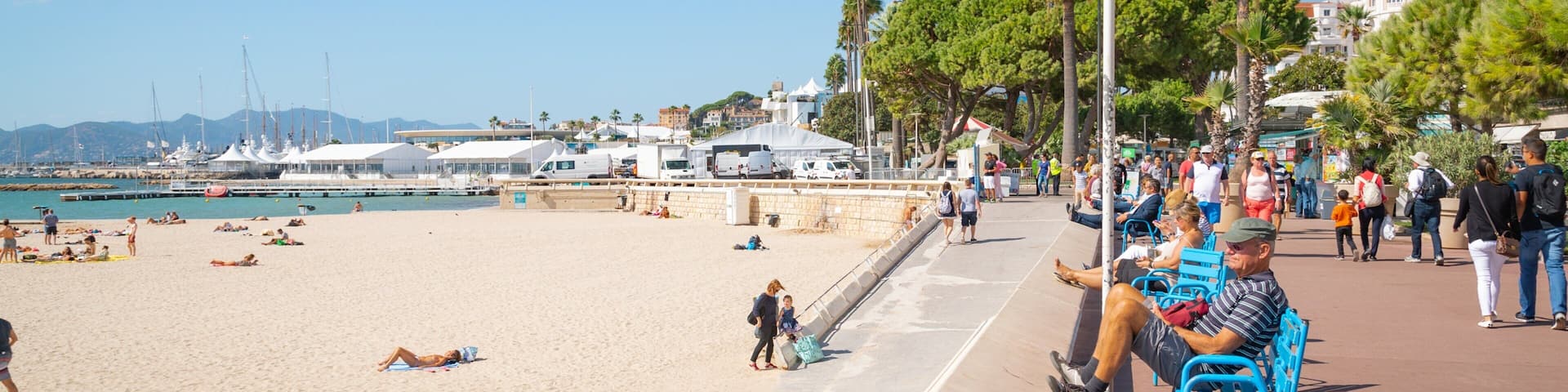 Una playa con gente relajándose, un paseo marítimo con bancos y un puerto deportivo con barcos.