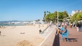 Una playa con gente relajándose, un paseo marítimo con bancos y un puerto deportivo con barcos.