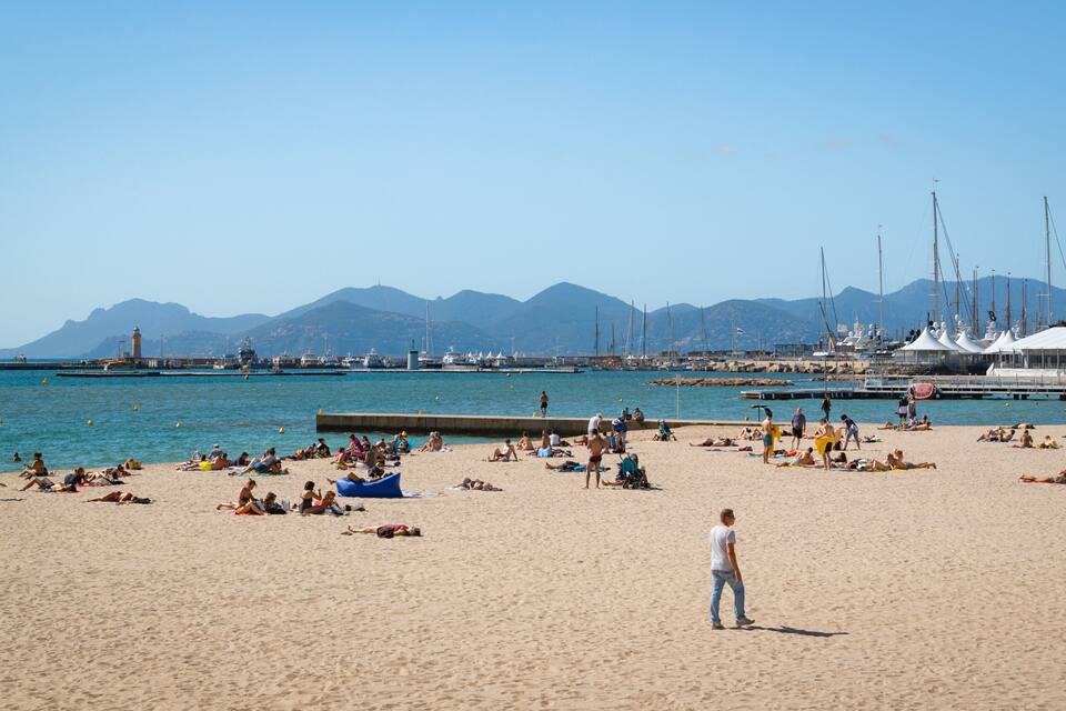 Een druk strand met mensen die zonnebaden en zwemmen, een vuurtoren en zeilboten op de achtergrond.