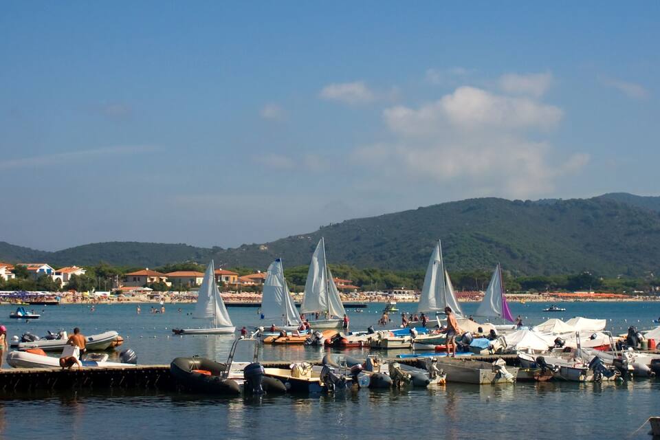 Ein Yachthafen mit Segelbooten, ein Strand und Berge im Hintergrund.