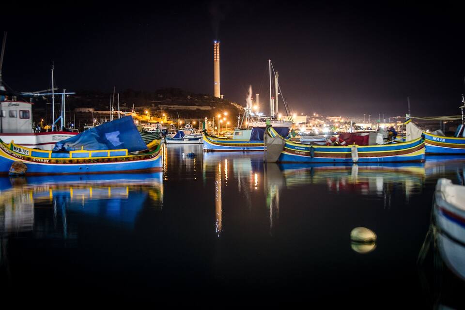 A nighttime harbor with several boats docked, reflecting lights on the water.
