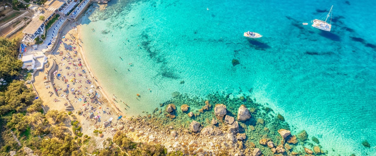A coastal area with clear turquoise waters, rocky outcrops, and a beach with umbrellas.