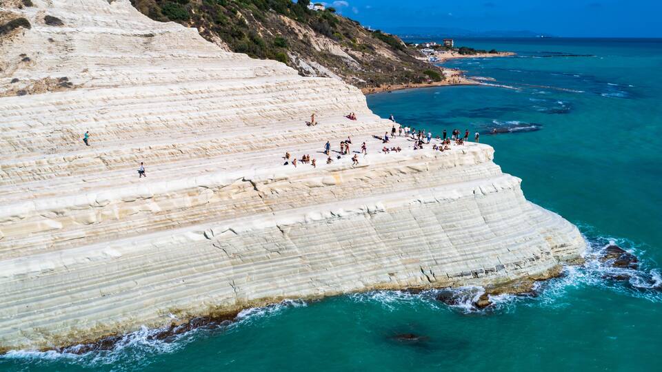 A coastal cliff with layered sedimentary rock formations and a beach with people.