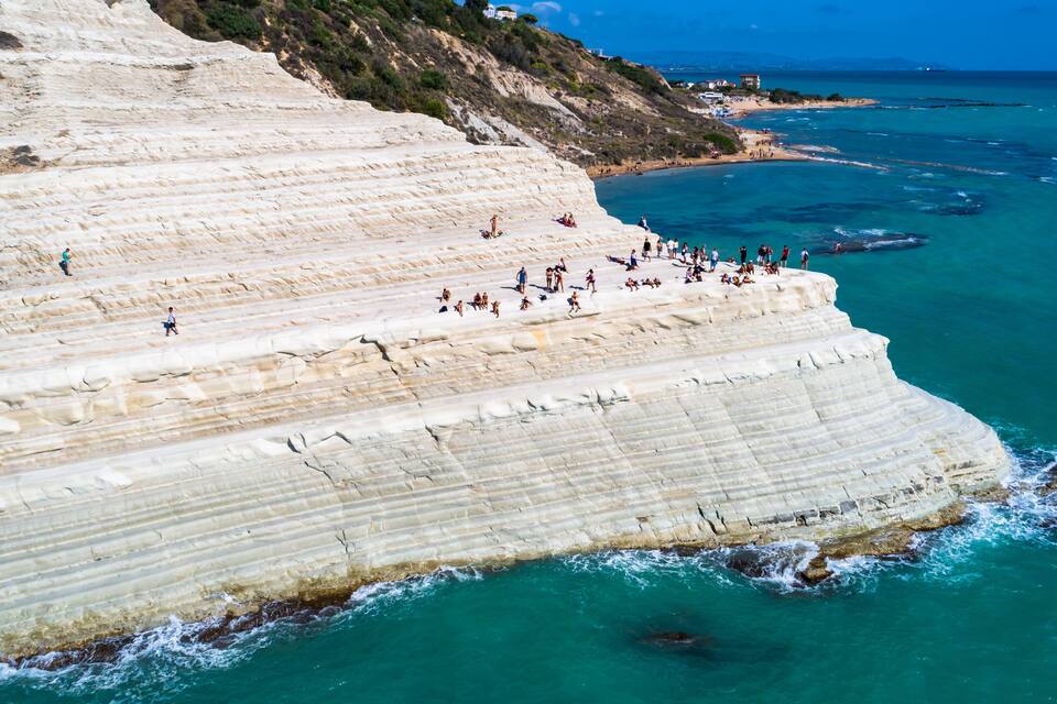 Una scogliera costiera con formazioni rocciose stratificate e una spiaggia con persone.