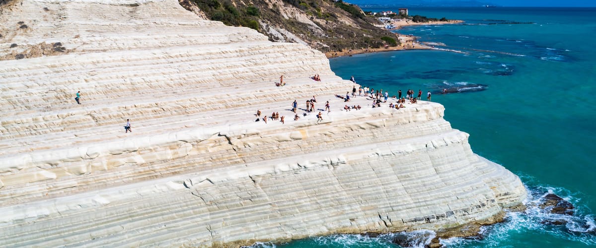 A coastal cliff with layered sedimentary rock formations and a beach with people.