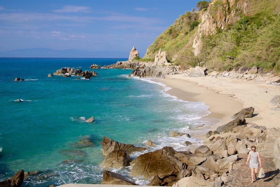 A rocky coastline with clear turquoise waters, a sandy beach, and a grassy hillside.