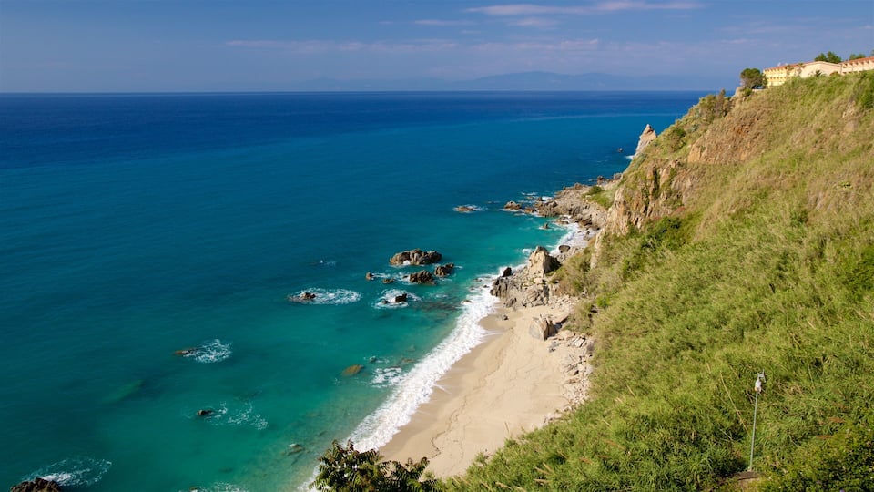 A coastal landscape with a sandy beach, clear turquoise waters, rocky outcrops, and a grassy hillside with buildings in the background.