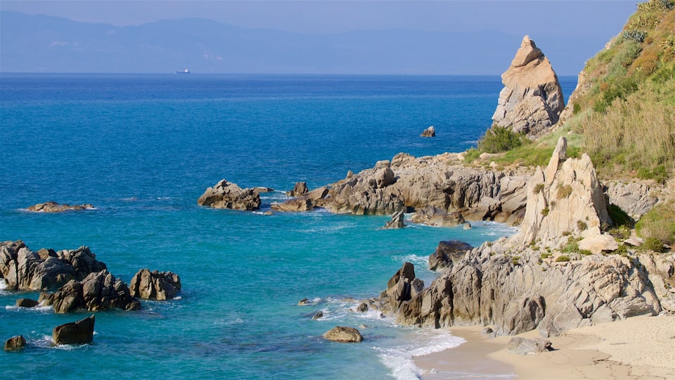 A coastal landscape with rocky outcrops, clear blue sea, and distant mountains.