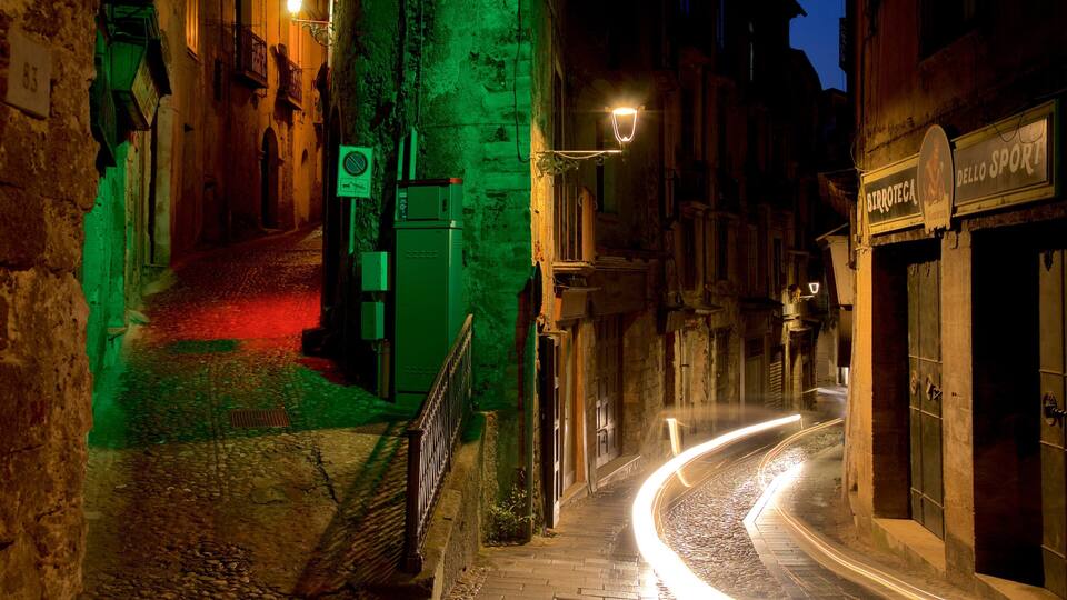 Ruas de paralelepípedos estreitas à noite, com letreiros iluminados e um rastro de luz deixado por um veículo em movimento.