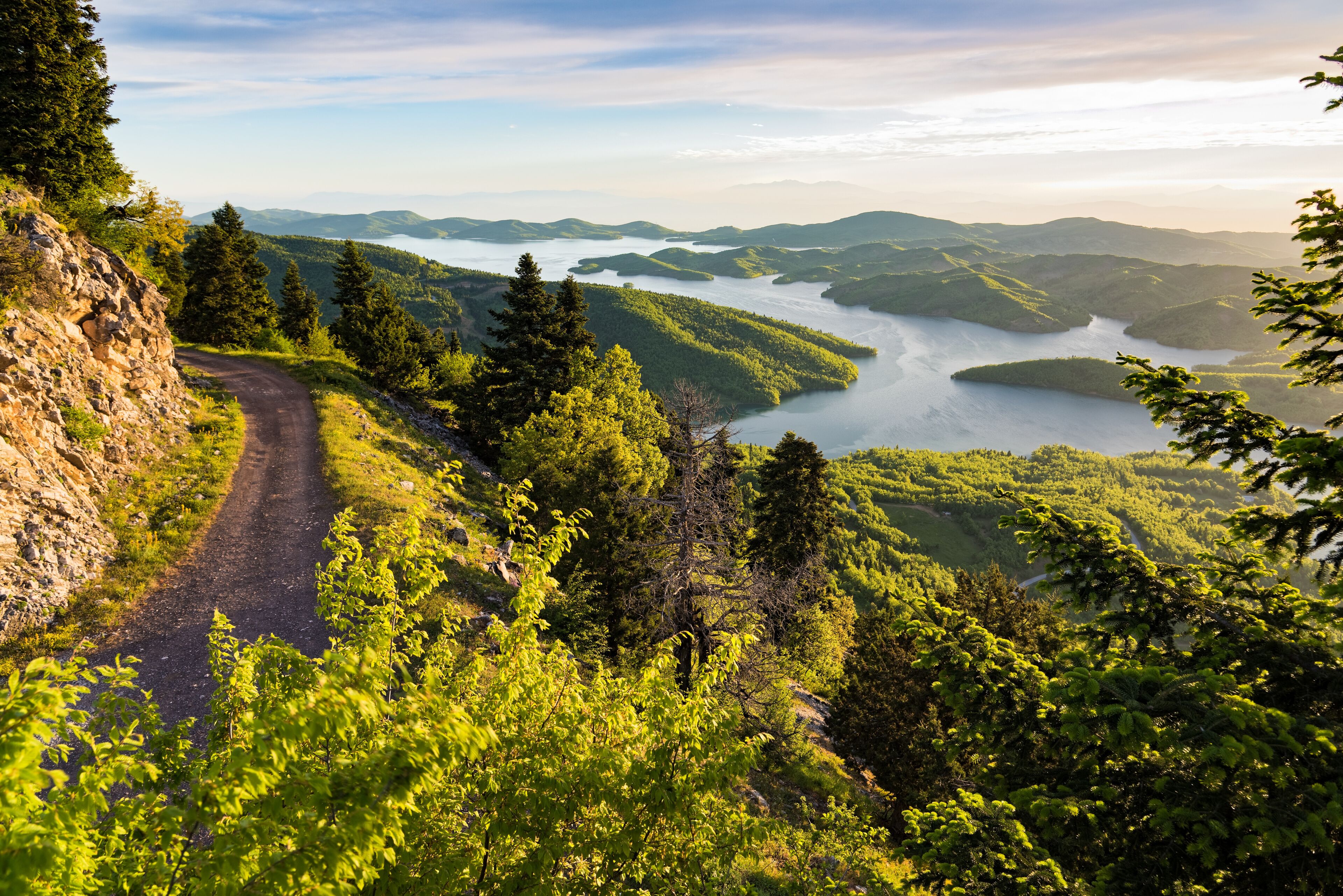A winding mountain road, dense forest, and a lake surrounded by hills.