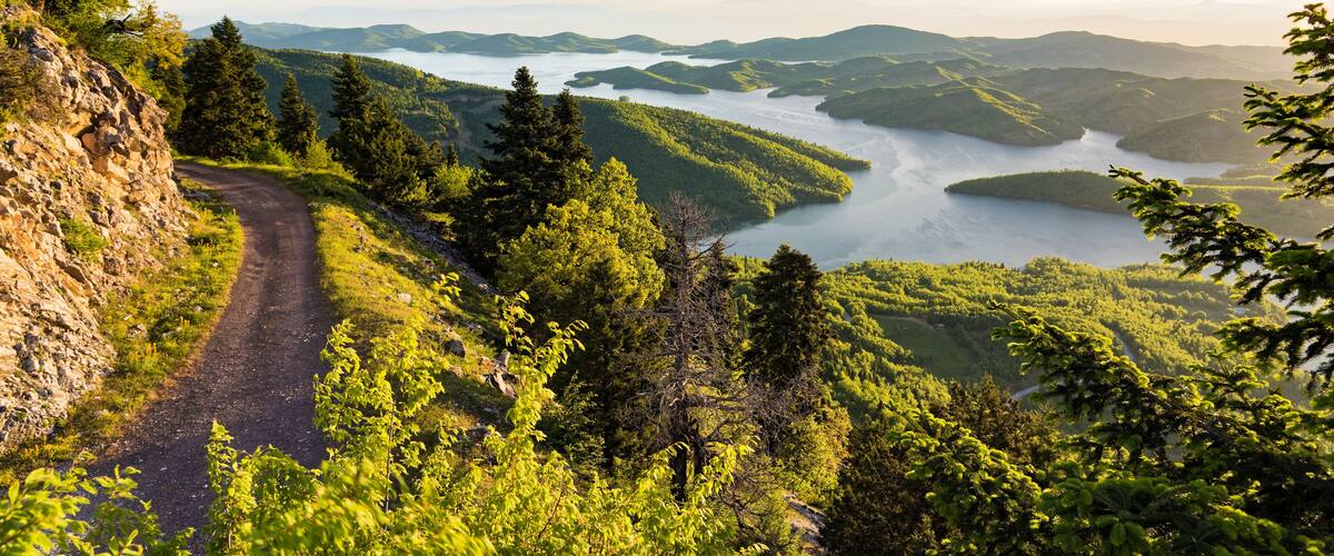 A winding mountain road, dense forest, and a lake surrounded by hills.