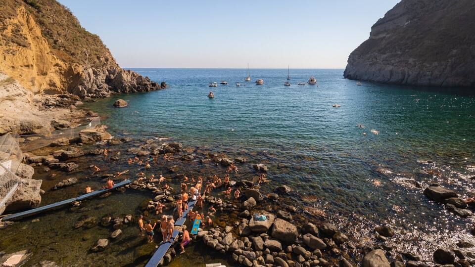 A rocky beach with clear water, people swimming and sunbathing, and boats in the distance.