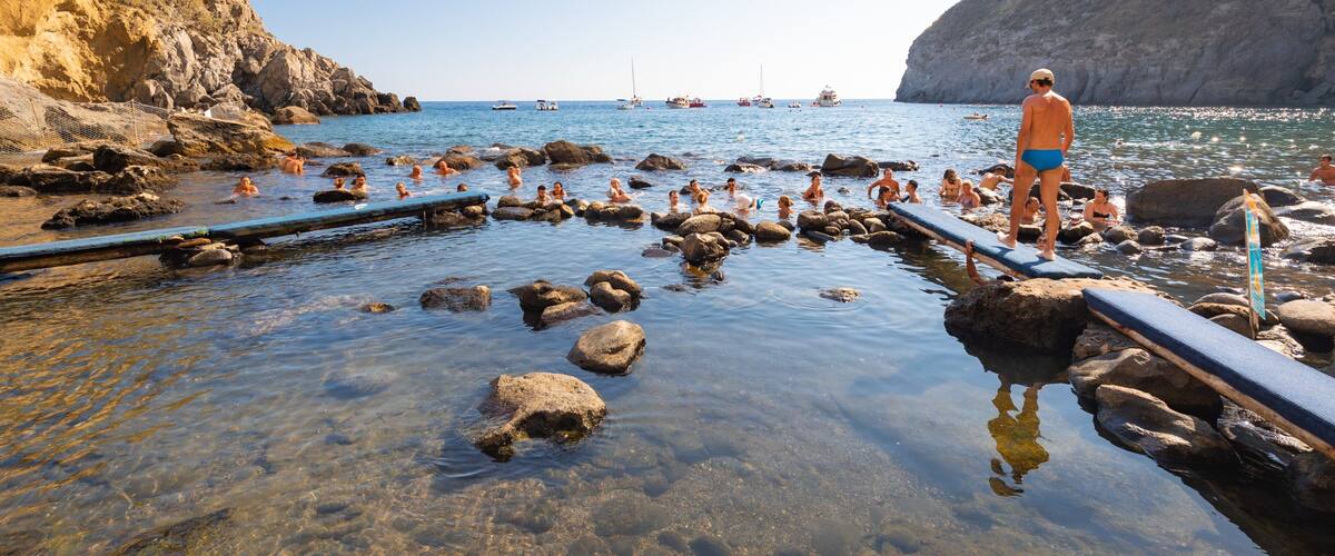 A rocky beach with clear water, people swimming, and boats in the distance.