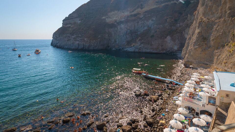 A rocky beach with clear turquoise water, boats, and a cliff.