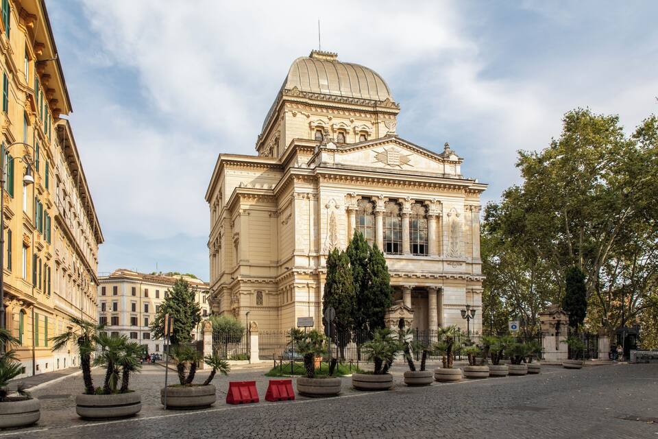 A historic building with a dome and columns, surrounded by a cobblestone square and trees.