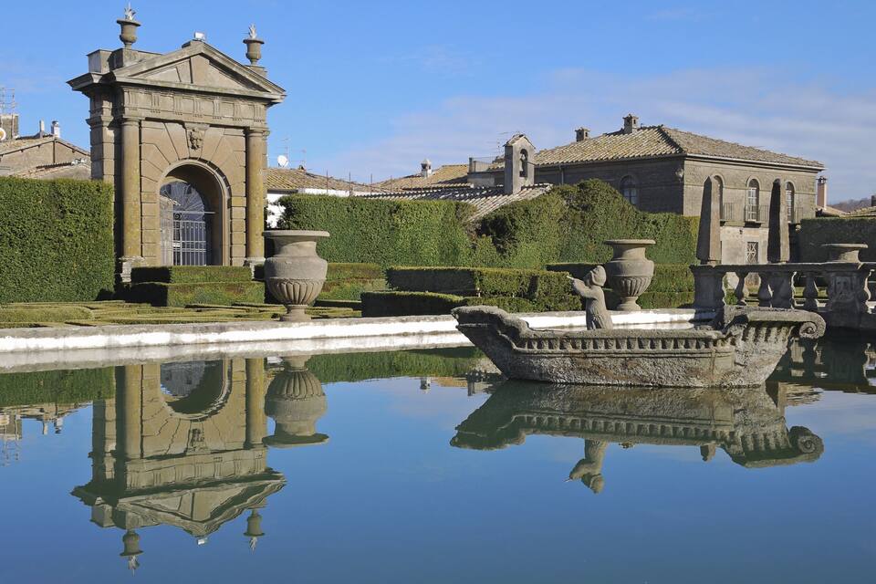 A classical garden with a reflective pond, a stone bridge, and a building with a tiled roof.