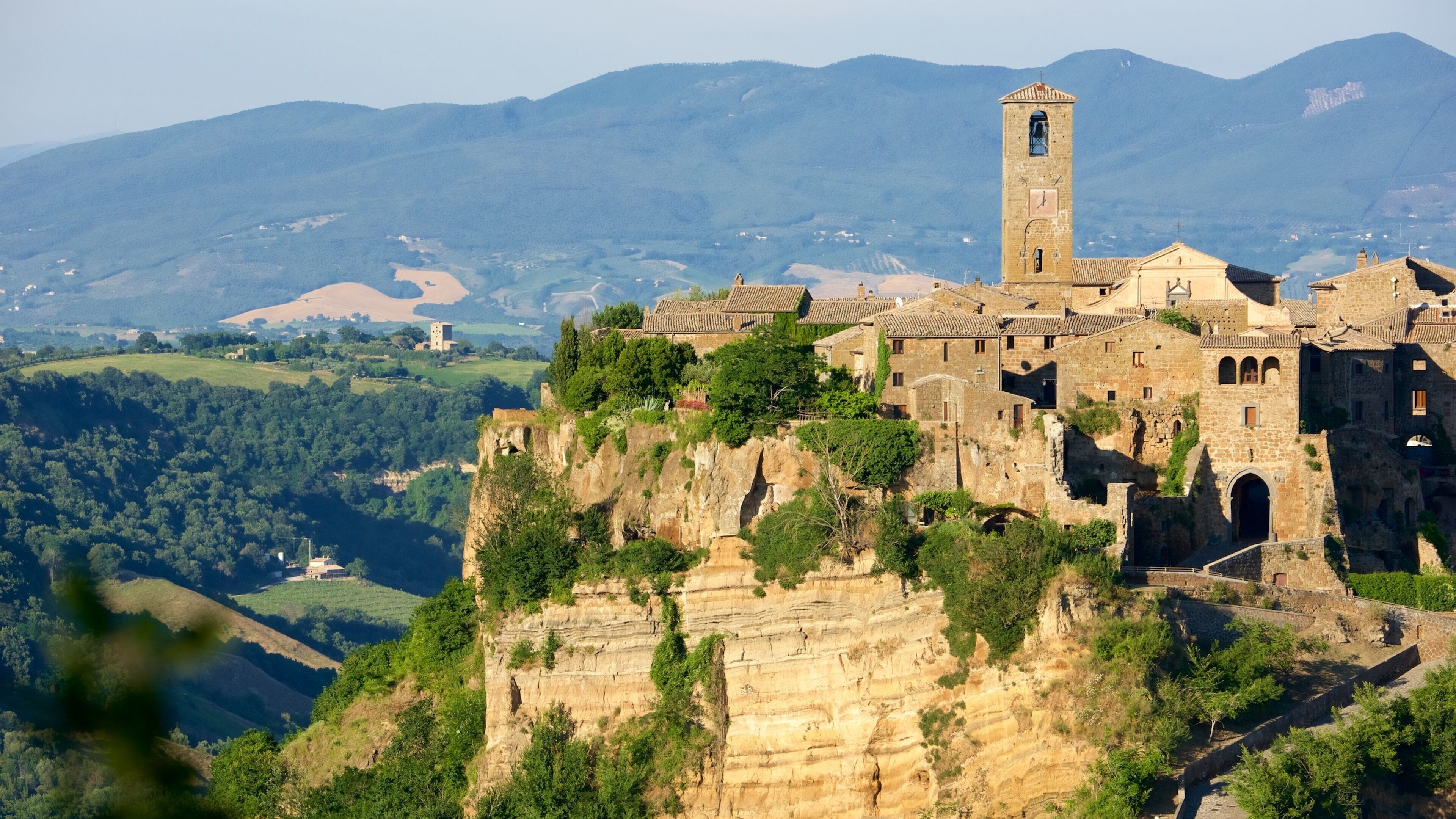 A historic stone building with a bell tower, perched on a cliff overlooking a valley with mountains in the background.